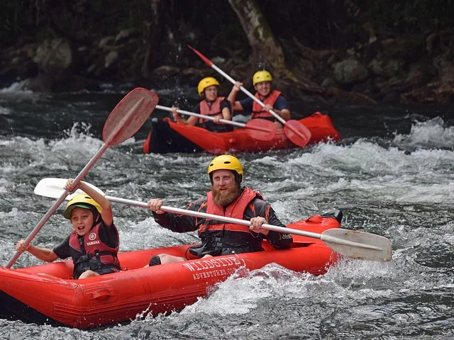 Mann und Kind in einem kleinen roten Schlauchboot, die auf dem Fluss paddeln, dahinter ein weiteres Raft.